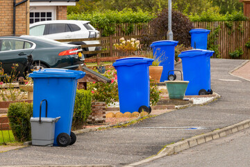 Blue waste recycling wheelie bins trash cans lined up in a row for collection on a residential street with parked cars in driveways