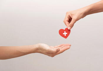 close up of Hand giving a red heart with a medical cross to another hand on grey background, organ...