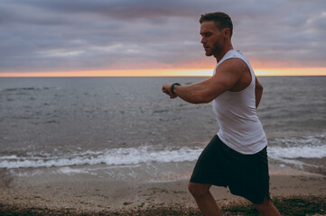 Side view young strong sporty athletic toned fit sportsman man wear sports clothes look at hand smart watch warm up train at sunrise sun dawn over sea beach outdoor on seaside in summer day morning