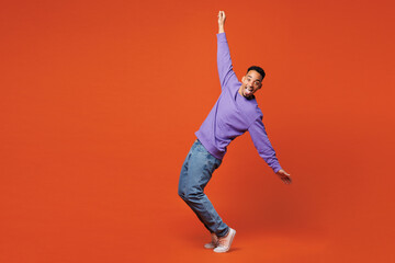 Full body young man of African American ethnicity wear purple sweatshirt casual clothes stand on toes dancing lean back have fun spread hands isolated on plain red orange background Lifestyle concept