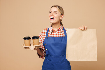 Young woman barista bartender barman employee she wears blue apron red shirt work in coffee shop hold paper takeaway bag, cup of tea isolated on plain beige background. Small business startup concept.