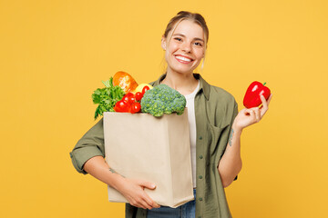 Young smiling woman wear casual clothes hold bag for takeaway mock up with food products show red bell pepper look camera isolated on plain yellow background. Delivery service from shop or restaurant.