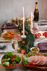 Christmas feast table with plates of food, glasses of wine and champagne, with festive decorations of pinecones, golden stars paper garland and long candles in evergreen foliage centerpieces