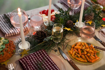 Christmas feast table with plates of food, glasses of wine and champagne, with festive decorations of pinecones, golden stars paper garland and long candles in evergreen foliage centerpieces