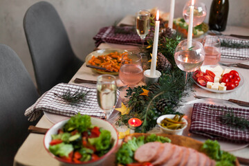 Christmas feast table with plates of food, glasses of wine and champagne, with festive decorations of pinecones, golden stars paper garland and long candles in evergreen foliage centerpieces