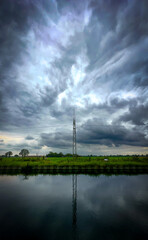 A striking view of a tall communication tower set against a stormy sky, beautifully reflected in the calm water below