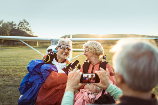 Two senior women laughing after skydiving, captured on camera by a friend