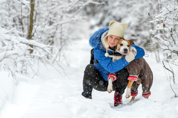 Young caucasian woman in blue jacket and black tights is sitting in the snowy forest and hugging his cute American Staffordshire terrier dog in a warm coat. Extremely cold weather. Kiisa, Estonia.