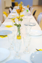 A served table with a white tablecloth, empty dishes and yellow flowers in vases. Beautiful serving and serving of dishes at the banquet. Catering.