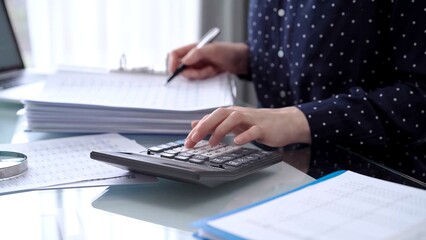 Businesswoman using calculator and reviewing ring folder of financial documents with magnifying glass at desk in modern office. Audit and taxes in business
