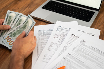 Fototapeta premium Hands counting hundred-dollar bills with tax forms and a laptop on a wooden table. Financial management and tax filing concept.
