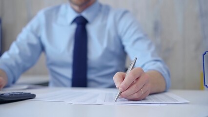 Unknown man accountant with blue t-shirt concentrating while calculating costs and taking notes at his desk. Taxes, audit in business