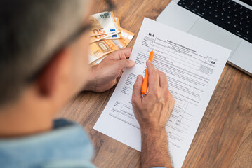 Close-up of a man filling out a tax form with an orange pen next to a stack of euro banknotes. Tax...