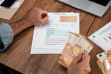 Obraz premium Man analyzing utility bill while holding euro banknotes at home. Focus on household expenses, tax management, and online payment solutions 