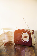 A retro-style radio is on a wooden table next to porcelain white mugs, illuminated by warm sunlight. Morning tea. Vertical photo.