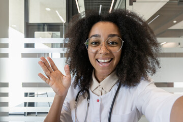 Close-up photo of a smiling young African American female doctor talking to the camera, waving and...