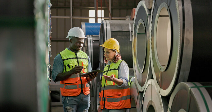 African American engineer man discussing women worker in construction warehouse steel plant factory. Engineer wear safety reflective vest white hard hat use smart tablet hand point checking product