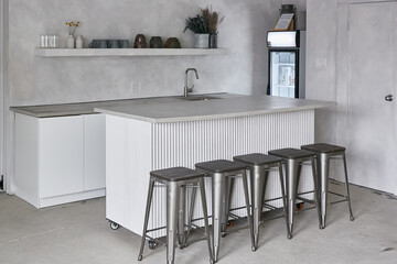 A minimalistic and modern kitchen setup with a sleek  countertop, industrial metal bar stools, and soft gray concrete walls. The kitchen features clean lines, a simple faucet, and open shelving 