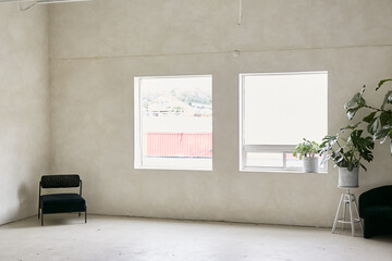 A minimalist room with large windows, neutral-toned walls, natural light, and a serene atmosphere. Features a dark chair in the corner, indoor plants, and an open, uncluttered layout.