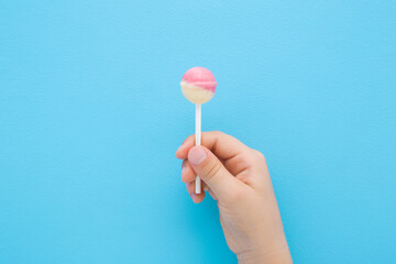 Little child fingers holding white pink lollipop on stick on light blue table background. Pastel color. Sweet candy. Closeup. Top down view.
