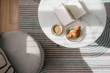 Cup of coffee, croissant and a book on a marble table close to armchair in living room