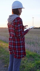 Woman engineer wearing a white protective helmet is taking notes with a clipboard in a field with wind turbines, as the sun sets. Clean energy and engineering concept