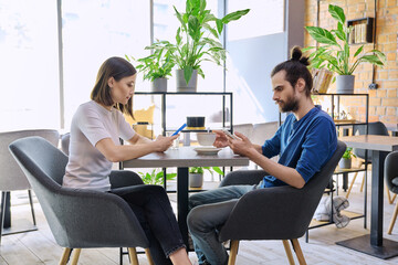 Fototapeta premium Young couple sitting at table in coffee shop, using smartphones