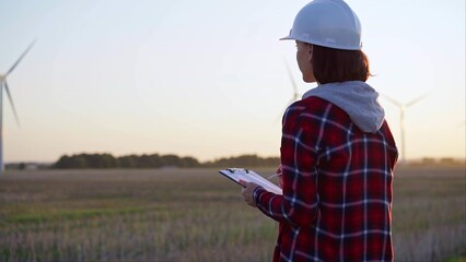 Adult woman engineer wearing white cask is taking notes on a clipboard on a field with wind turbines, as the sun sets, back view