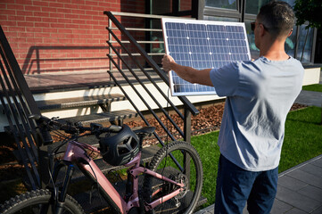 Back view of man holding solar panel toward the sun for charging electric mountain bike. Pink...