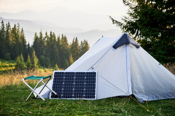 White tent equipped with solar panel sits in grassy field surrounded by lush trees and hills. Solar...