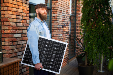 Portrait of bearded man holds solar panel, standing in front of brick building and lush greenery....