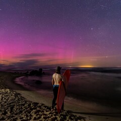 surfer with board on beach at night under starry sky and aurora borealis.