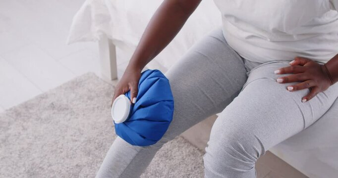 Close-up of an African American woman applying an ice pack to injured knee while massaging the leg. The cold treatment helps relieve pain and ache from the injury, providing hurt relief at home.
