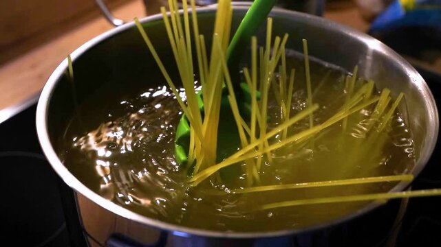 Spaghetti being added to boiling water on a kitchen stove and being stirred by a green  spoon. Cooking pasta for a homemade Italian dinner in a stainless steel pot. Traditional home culinary. 4K video