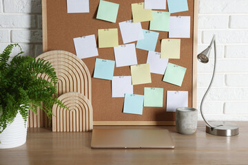 Obraz premium Cork board with blank paper notes, laptop and decorative elements on wooden table near white brick wall