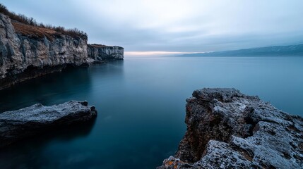 Serene coastal view at dawn with smooth water and rocky formations under a cloudy sky.
