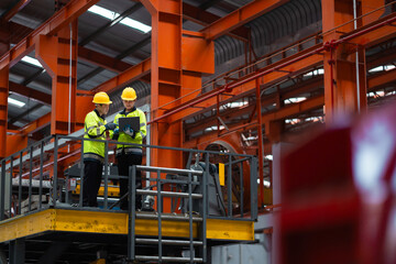 Two men are standing on a platform in a factory, looking at a computer screen. They are wearing yellow and black safety gear