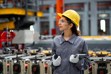 A woman wearing a yellow helmet and safety glasses stands in a factory. She is holding a tablet in her hand