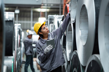 A woman wearing a hard hat and safety glasses is looking at a tablet while standing in a warehouse