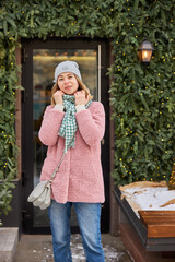 Young woman in a cozy pink jacket and scarf stands outside a festive winter cafe adorned with greenery and lights during the holiday season