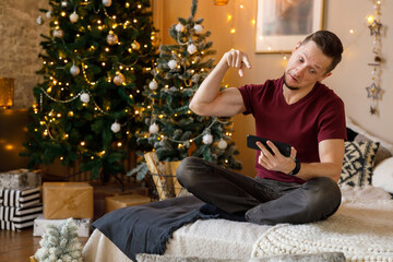 man sitting cross-legged on a cozy bed, swears a video call amidst festive Christmas decorations in a warm living room