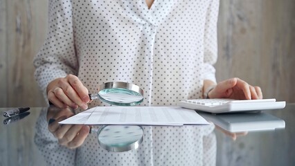 Auditor woman analyzing financial documents with magnifying glass and calculator at her office desk in formal white blouse with polka dots. Business people concept
