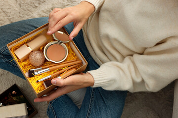 Woman holding Christmas gift box with makeup brushes and cosmetics at home, above view