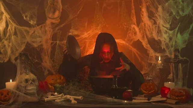 A witch in black clothes is cooking a potion in a scary kitchen with pumpkins and a human skull and bones.