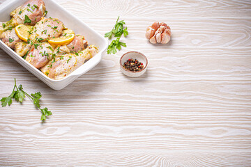 Raw uncooked chicken legs with seasonings, herbs, lemon in white ceramic casserole top view on light wooden rustic background. Preparing healthy meal with marinated chicken drumsticks. Space for text.