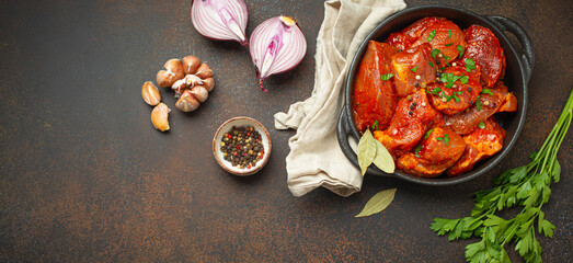 Cooking hearty meat stew. Raw uncooked chopped pieces of meat marinated with seasonings and parsley in black casserole dish top view, ingredients on dark rustic background. Space for text.