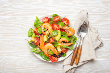 Healthy salad with grilled shrimps, avocado, cherry tomatoes and green leaves on white plate with cutlery on white wooden rustic background top view. Clean eating, nutrition and dieting concept..