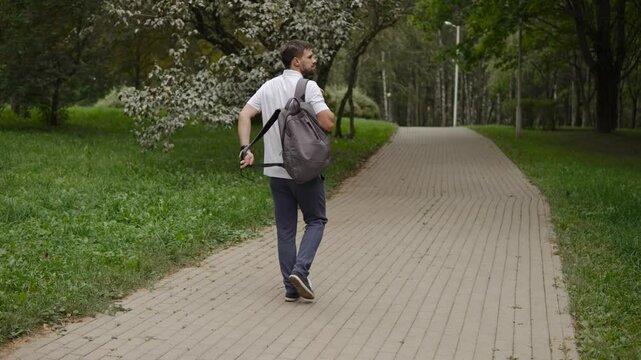 Caucasian man puts backpack on back while walking in park, back view. Walking on tiled sidewalk among trees in city park with definite purpose. Cloudy windless summer day.