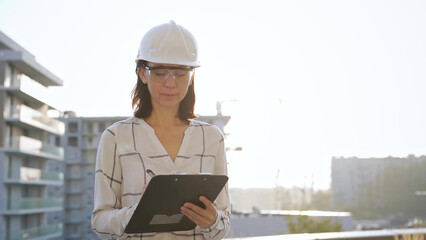 Woman construction engineer wearing white checked blouse and hard hat is making notes on a clipboard while inspecting a building site at sunset, front view. Architecture and engineering concepts