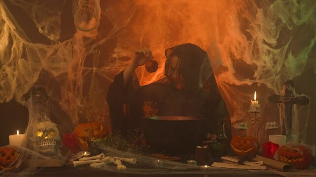 A witch in black clothing pulls a poisoned apple from a cauldron in a scary kitchen with pumpkins and human bones.